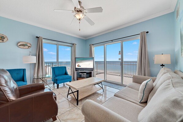 A living room with a sofa, armchair, and a coffee table. There is a ceiling fan, a TV, and a view of the ocean through large windows. - Crystal Shores West 101- Enjoy the View of Paradise (Gulf Shores)