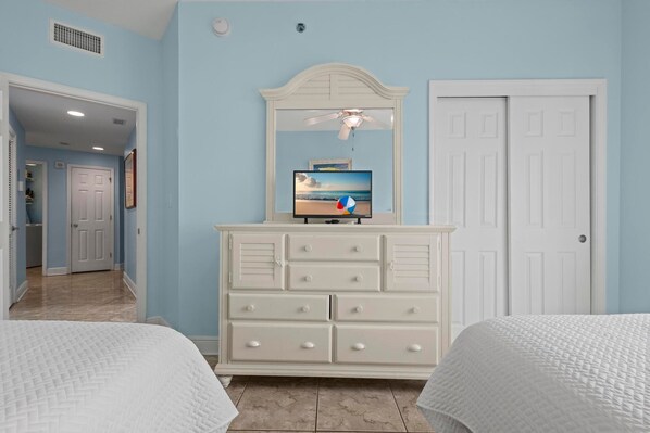Une chambre avec deux lits, une commode, un ventilateur de plafond et un téléviseur posé sur la commode.
