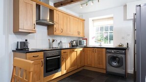 A modern kitchen with wooden cabinets, a stainless steel range hood, a microwave, a kettle, and a washing machine. - Pollys Cottage (Ambleside)