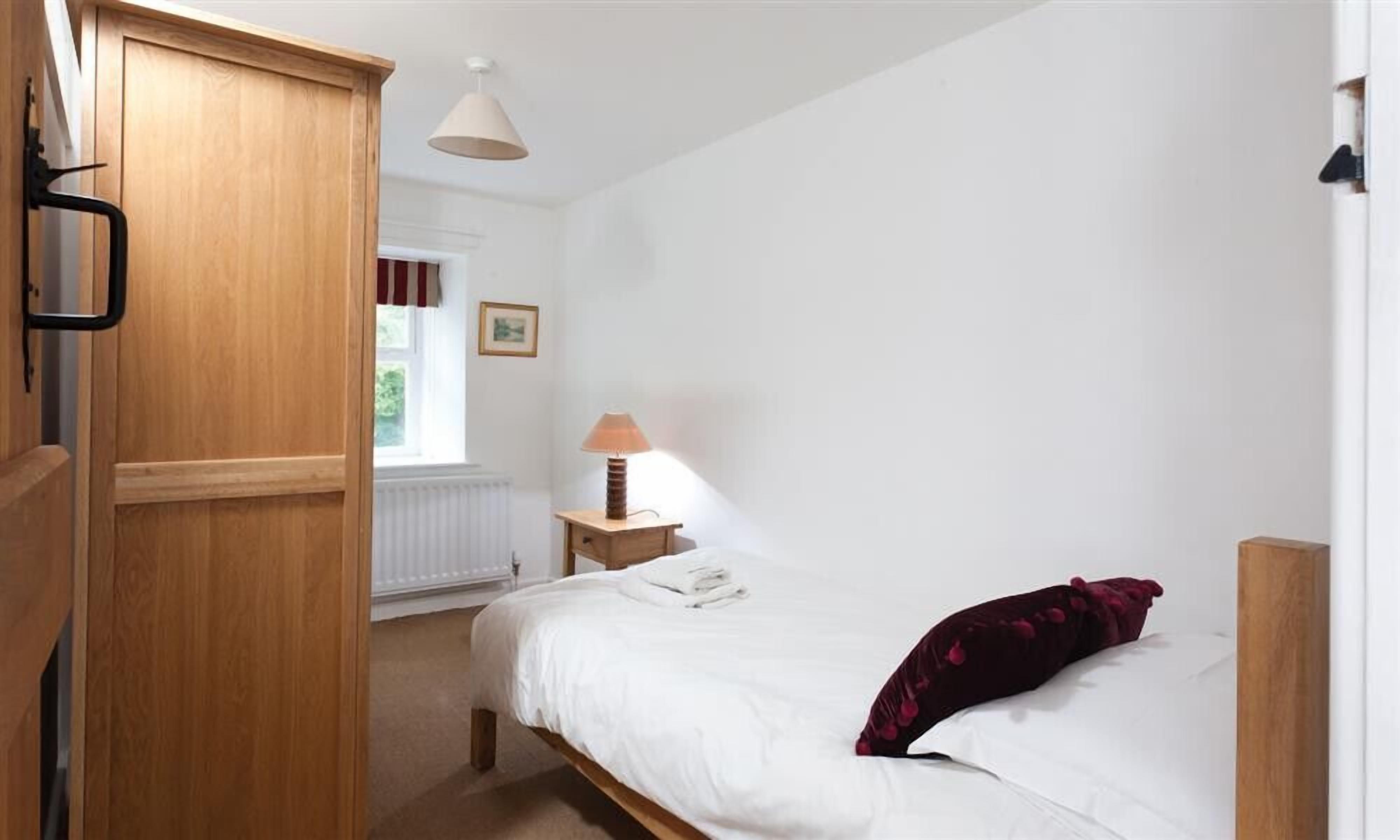 Une chambre avec une armoire en bois, une table de chevet avec une lampe, un lit avec une literie blanche et un oreiller rouge foncé, et une fenêtre avec des rideaux.