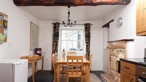 A compact kitchen with a dining area, featuring a wooden table and chairs, a chandelier, and a view through a window with patterned curtains. - Pollys Cottage (Ambleside)