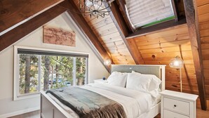 A bedroom with a sloped ceiling, a bed with white linens, a nightstand, a lamp, and a window with a view of trees. - Cobalt by Tahoe Getaways - Luxury 3BR w/ Hot Tub, Walk to Lake - Dog OK (Carnelian Bay)