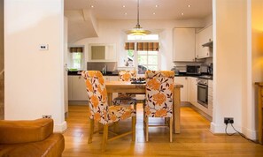 A modern kitchen with wooden cabinets, a dining table with floral chairs, and a built-in oven. - Rothay Holme Cottage (Ambleside)