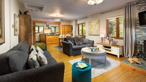 A modern living room with a sofa, a coffee table, and a fireplace. There is a kitchen area in the background with wooden cabinets and a dining table. - Brooklands (Ambleside)