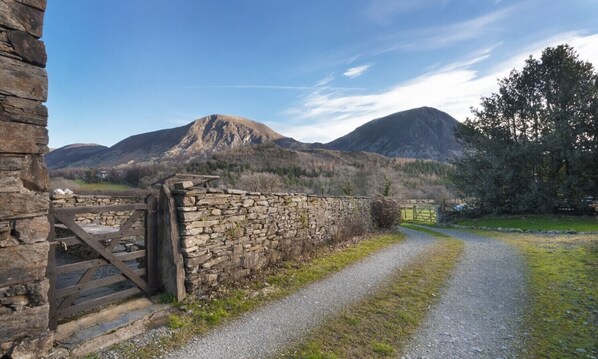 Un mur de pierres et un portail en bois mènent à un chemin de gravier dans une zone rurale, avec des montagnes en arrière-plan.