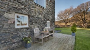 A stone building with a patio area featuring a table and chairs, a small potted plant, and a view of a grassy yard with trees. - Cedar Nook (Cockermouth)
