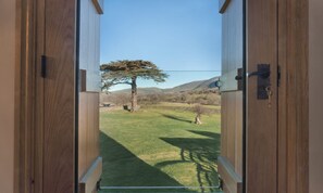 Une porte en bois donne sur un espace extérieur verdoyant, avec un arbre et des montagnes en arrière-plan.