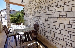 Een terras buiten, met een stenen muur, een tafel gedekt met een witte tafelkleed en stoelen, en een helder uitzicht op de straat en het groen op de achtergrond.
