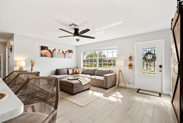 A spacious living room with a large window, a ceiling fan, and a dining area with wicker chairs.
