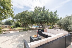 An outdoor patio with a sofa set, a table with food and drink, and a view of a swimming pool and trees.