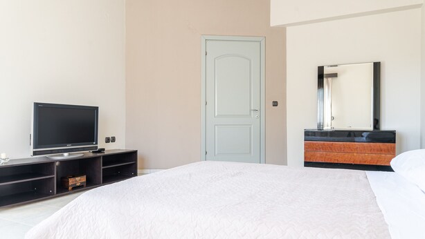 A hotel room with a bed, a TV on a stand, a door, and a vanity with a mirror. - Villa Archodia - With Private Pool (Prines)