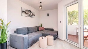 Ein modernes Wohnzimmer mit einem grauen Ecksofa, zwei runden Hocker, einer Stehlampe und einem großen Fenster mit Blick auf Grünflächen.