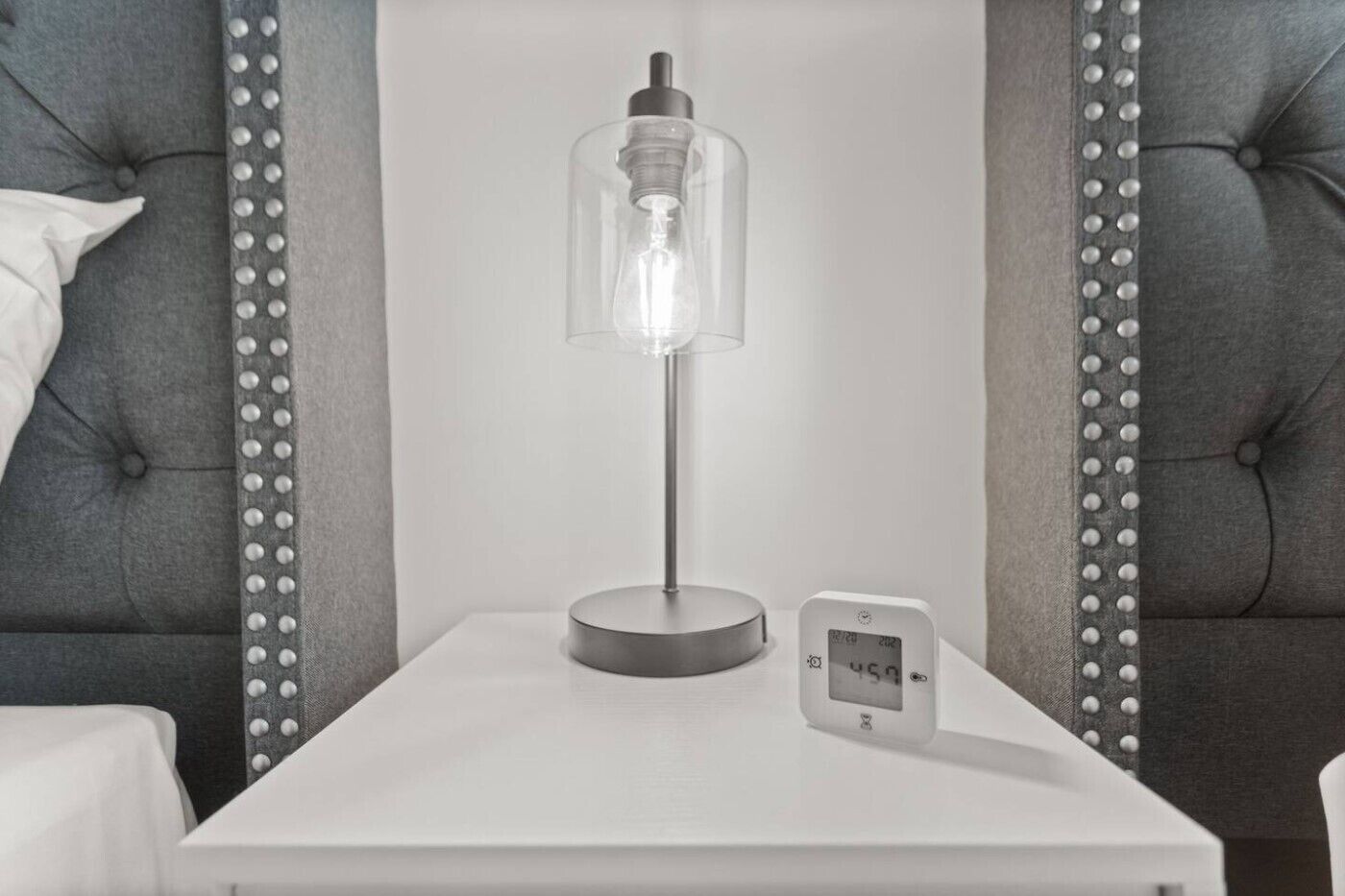 A bedside table with a clear glass lamp, a white bedside table, and a digital thermostat.