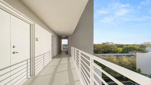 En gang med hvide døre, en balkon med gelænder og udsigt til træer og bygninger.