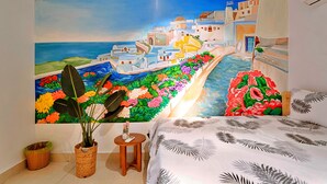 Une chambre à coucher ornée d’une fresque représentant une ville côtière, un lit recouvert d’une couvre-lit à motifs de feuilles, une petite table en bois et un panier en osier.