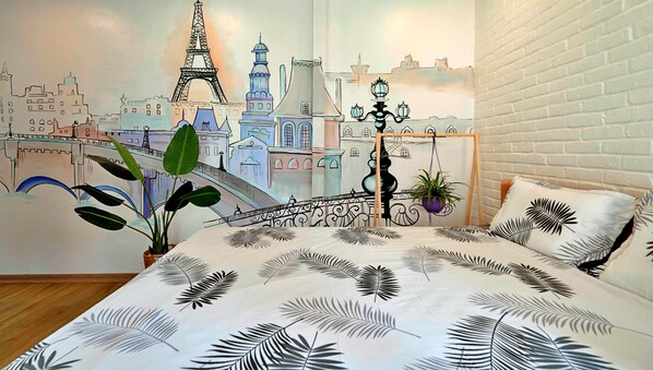 Un dormitorio con un mural de paisaje urbano, una cama con ropa de cama de hojas de palma, una planta en maceta y una lámpara colgante.