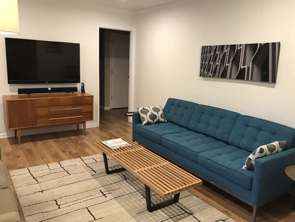 Un salon avec un canapé bleu, une table basse en bois, un téléviseur posé sur un meuble en bois et un tableau abstrait moderne accroché au mur.