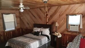 Une chambre confortable avec un plafond en bois, un lit recouvert d’une literie à motifs, une table de chevet avec une lampe et un fauteuil agrémenté d’un plaid à motifs.