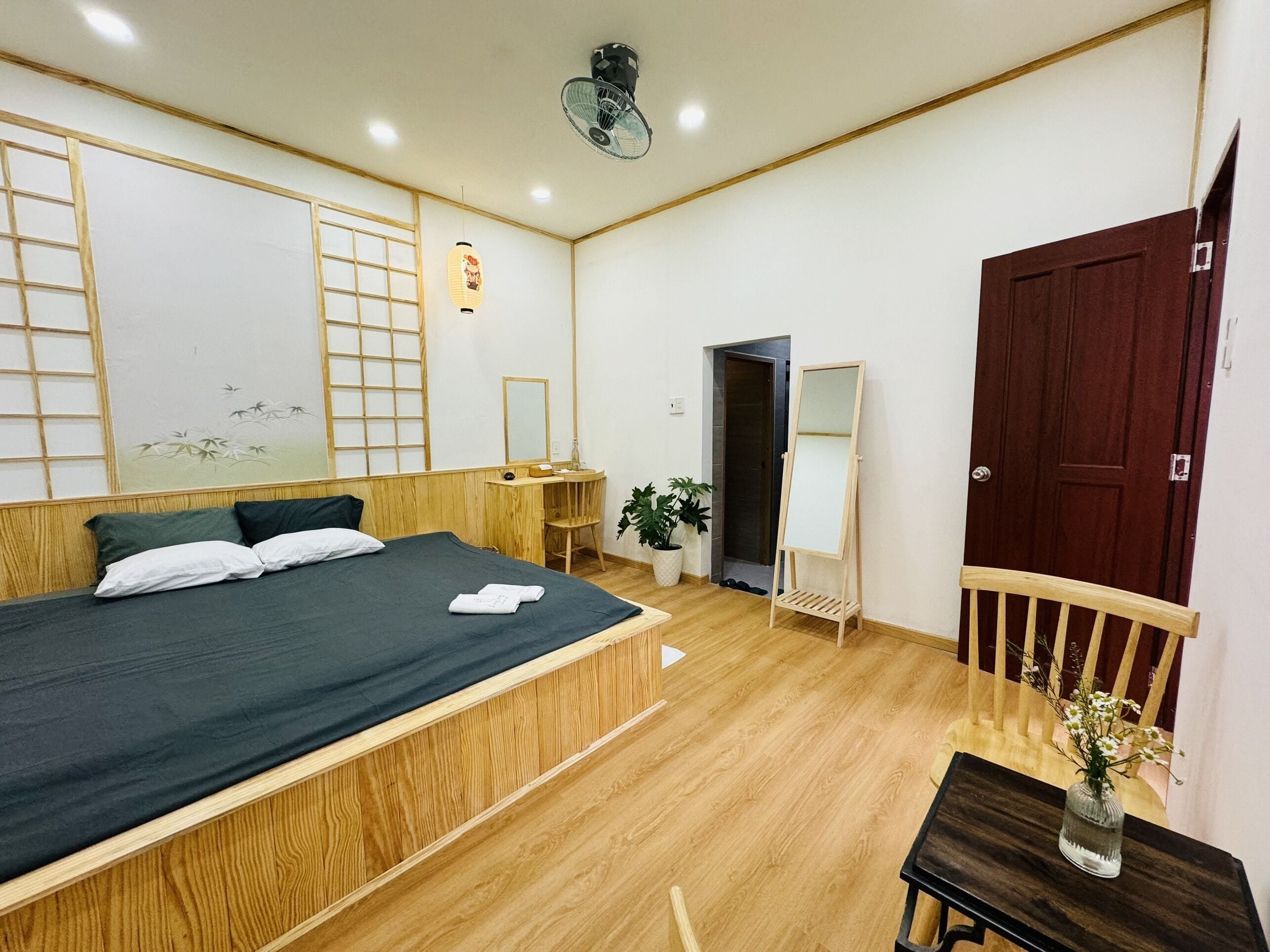 A hotel room with a large bed, wooden headboard, a small table with a vase, a mirror, and a wooden chair.