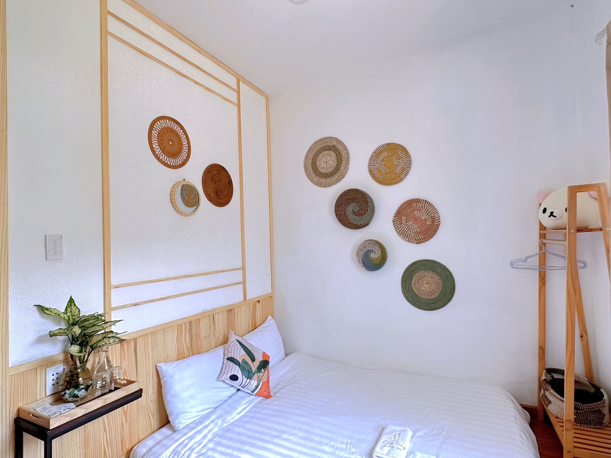 A neatly arranged bedroom with a bed, pillows, a bedside table with a vase, and a wall with circular woven decorations.