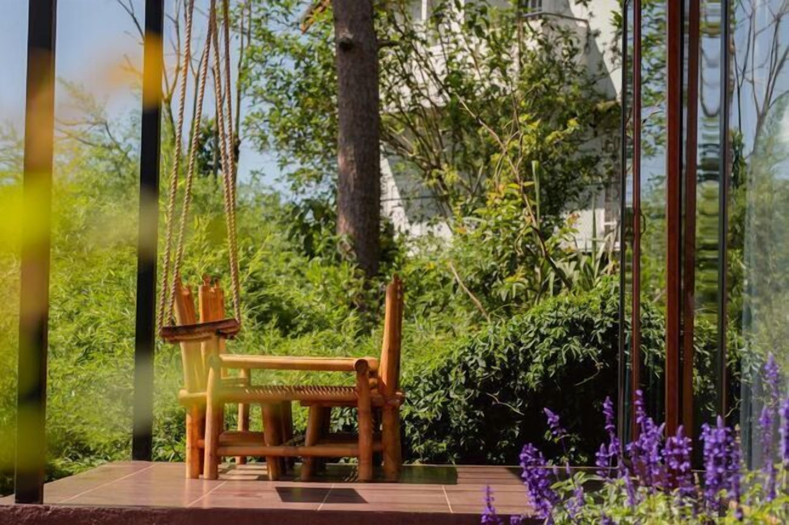 A wooden swing chair in a lush garden with dense foliage and a clear view of a building in the background.