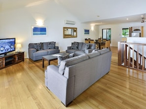 Ein modernes Wohnzimmer mit einem großen, grauen Ecksofa, einem Couchtisch aus Holz und einem Flachbildfernseher auf einem Holzständer. Es gibt einen Essbereich mit einem Holztisch und Stühlen sowie eine Küchenzeile mit einer Holztreppe.