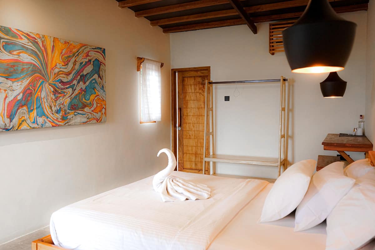 A neatly made bed with a swan-shaped towel, a colorful abstract painting on the wall, and a wooden ladder leaning against a door.