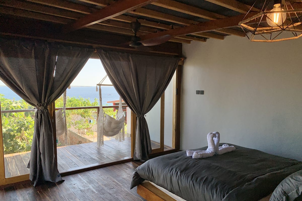 A bedroom with a bed, wooden floor, and a view of the sea through large windows.