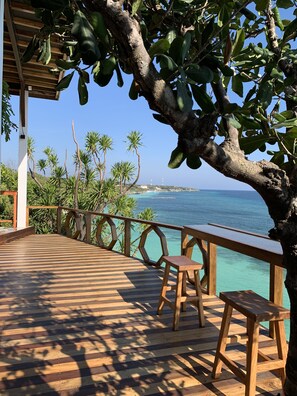 A wooden deck with a railing overlooking a clear blue sea, palm trees, and a distant view of a city skyline. - Seascapes Bira (Bira)