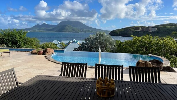 An infinity pool with a view of the sea and mountains, surrounded by outdoor furniture and greenery.