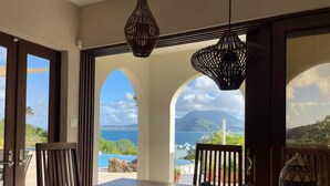 A dining area with a wooden table, two chairs, and a decorative bowl. There are two large windows with arched frames, offering a view of the sea and a mountain.