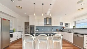 A modern kitchen with white cabinetry, a central island with bar stools, and a large window offering a view. - Luxury Mandalong Mansion with Pool & Spa (Mandalong)