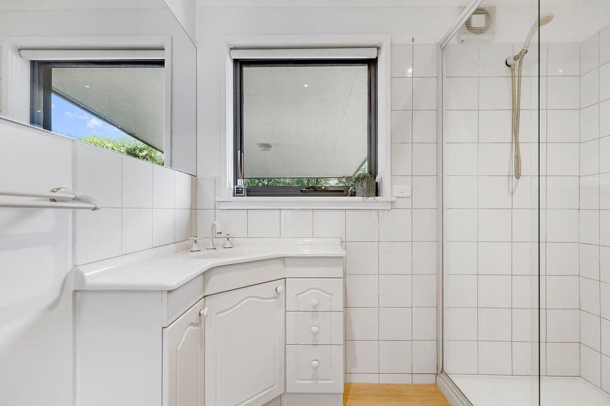 A modern bathroom with white tiles, a shower, a window with a view, and a sink with a mirror.
