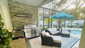 An outdoor kitchen area with a stone wall, a grill, and a poolside seating area with a blue umbrella.