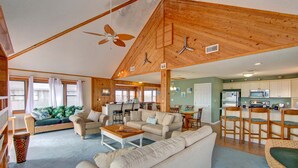 A spacious living area with a high ceiling, wooden beams, a large sofa, a coffee table, and a kitchen area with bar stools. - Semi-Oceanfront, Beach Access, Hot Tub & Pool Table (Avon)