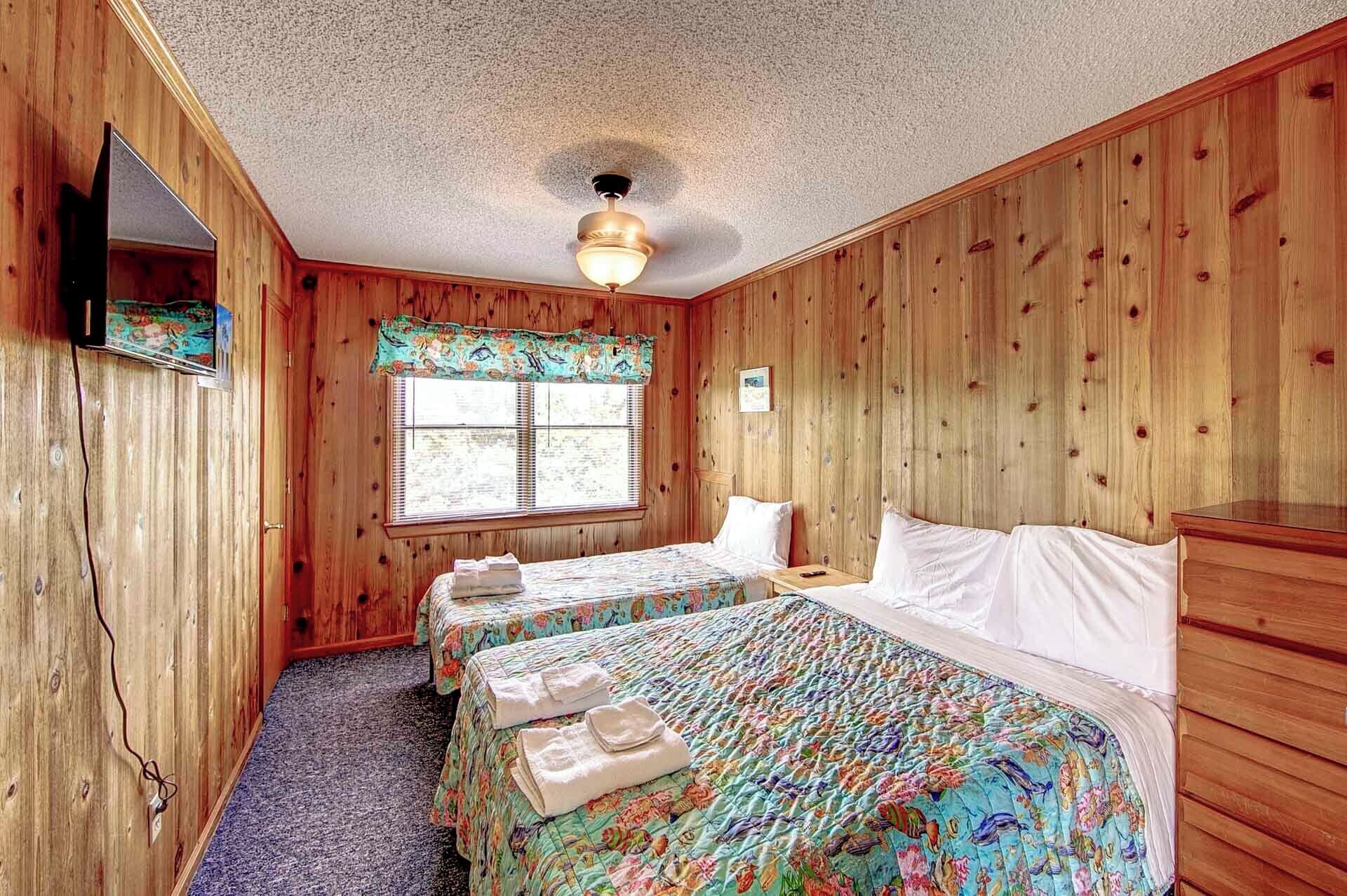 Une pièce aux murs en bois, avec un ventilateur au plafond, une fenêtre munie de stores, deux lits avec une literie à motifs floraux et une petite table avec une lampe.