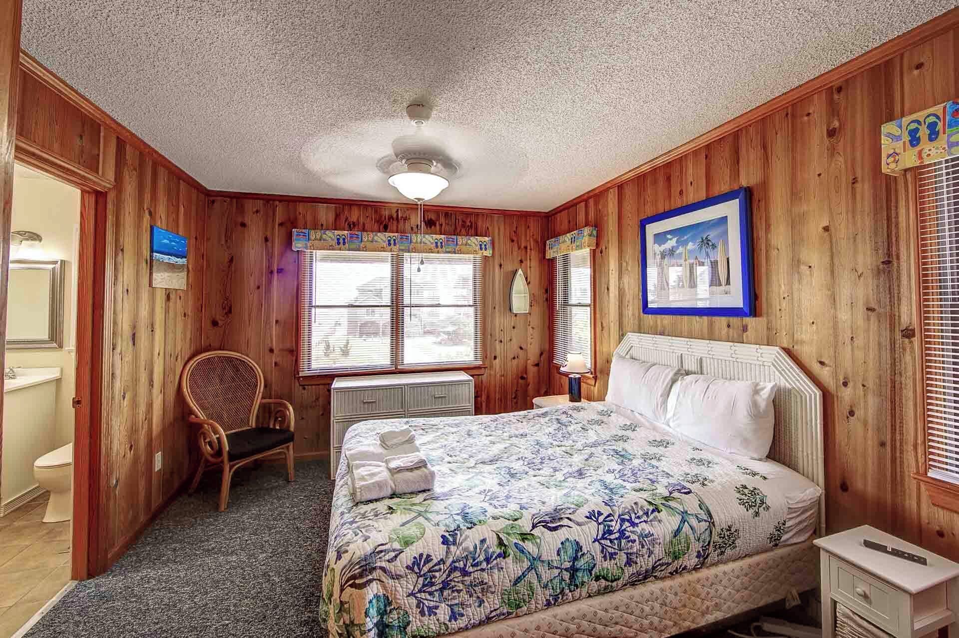 Une pièce aux murs en bois, un lit avec une literie à motifs floraux, une chaise, un bureau et un ventilateur de plafond.