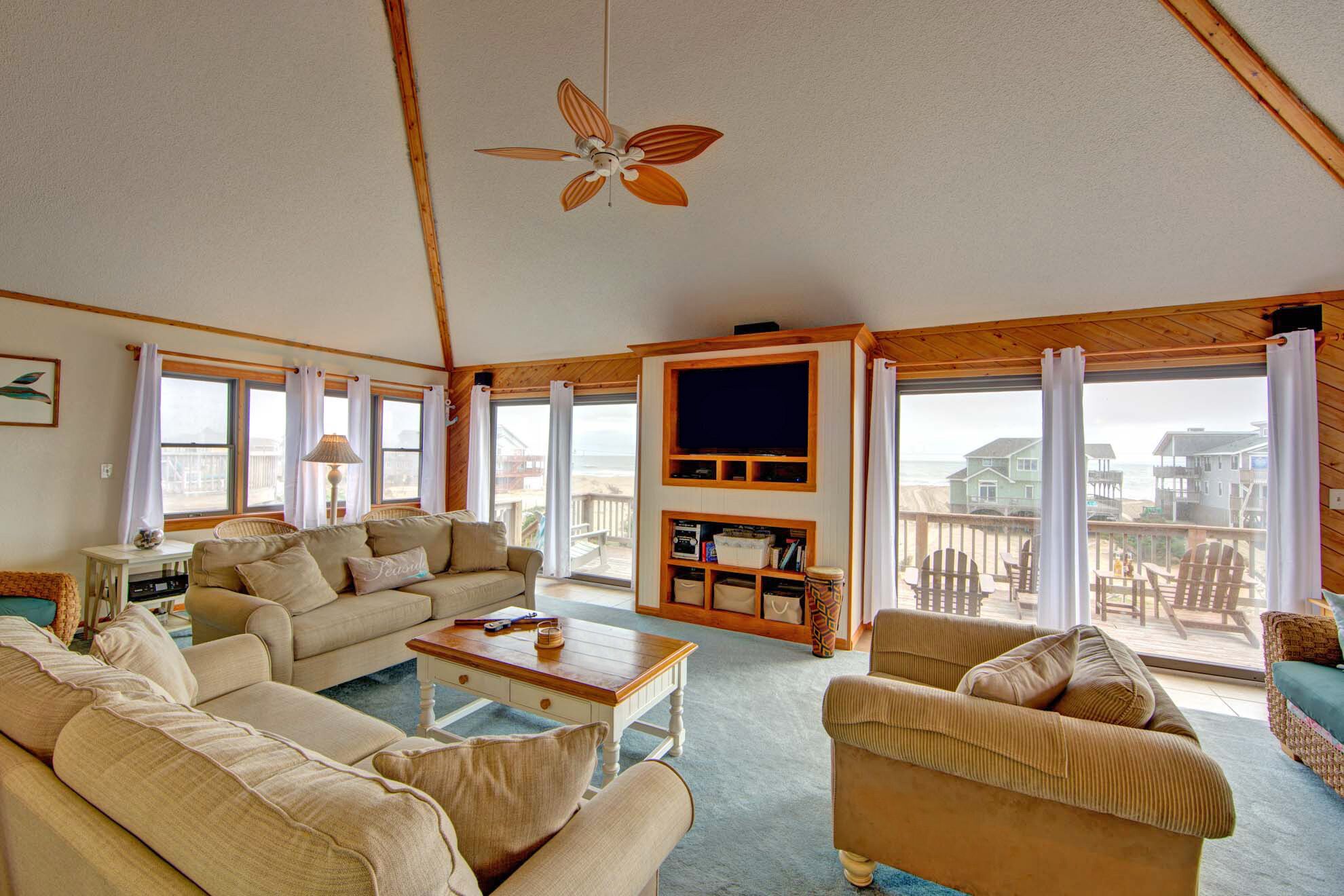 A spacious living room with a large TV, comfortable seating, and a balcony with a view of the beach.