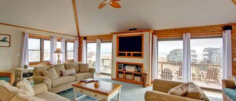A spacious living room with a large TV, comfortable seating, and a balcony with a view of the beach.