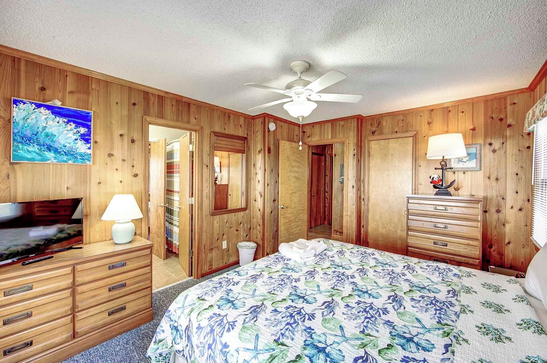 Un dormitorio con paneles de madera, una cama, un tocador, un televisor y un ventilador de techo.