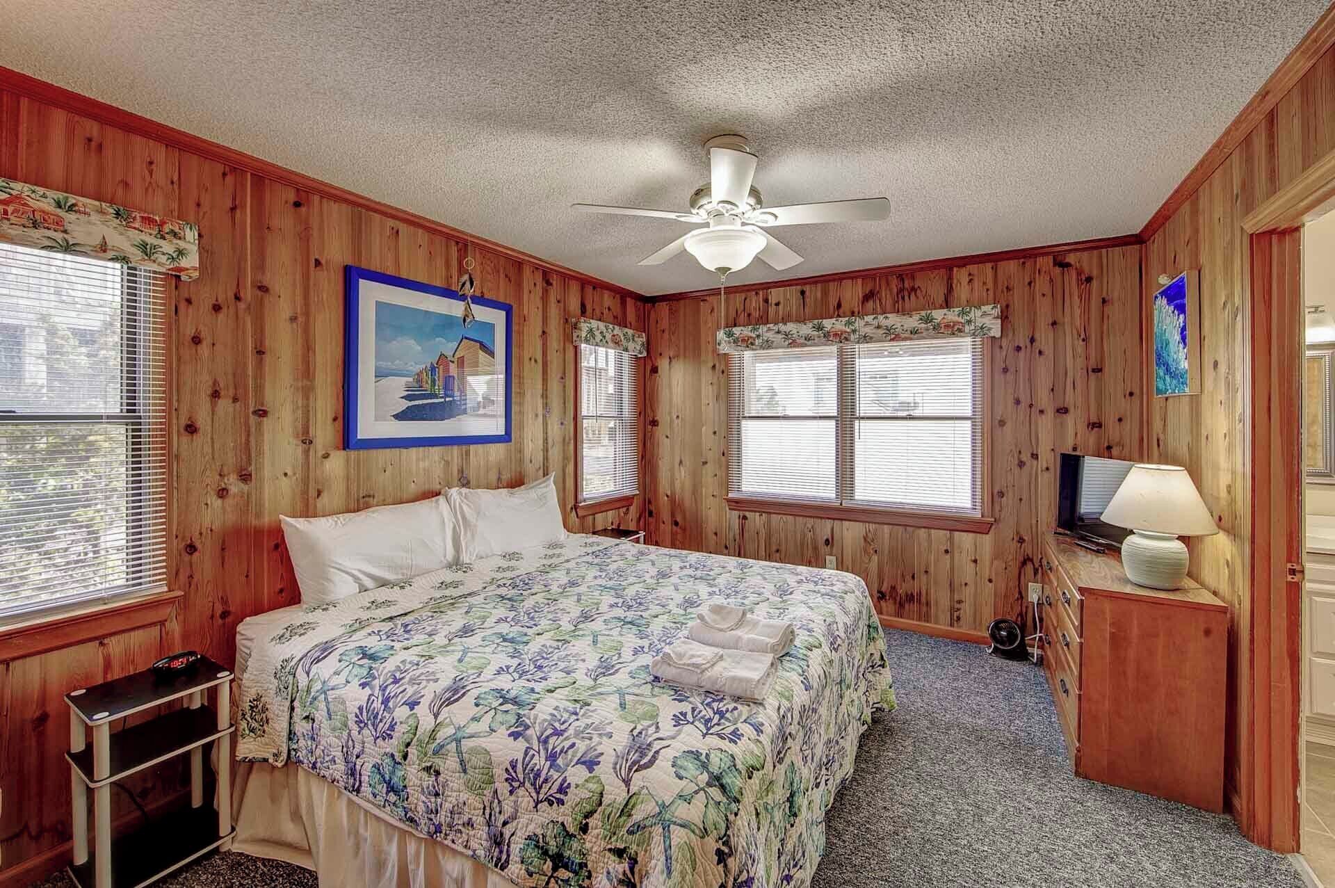 Une chambre avec des murs en bois, un lit recouvert d’une literie à motifs floraux, un ventilateur de plafond et un tableau représentant une scène de plage.