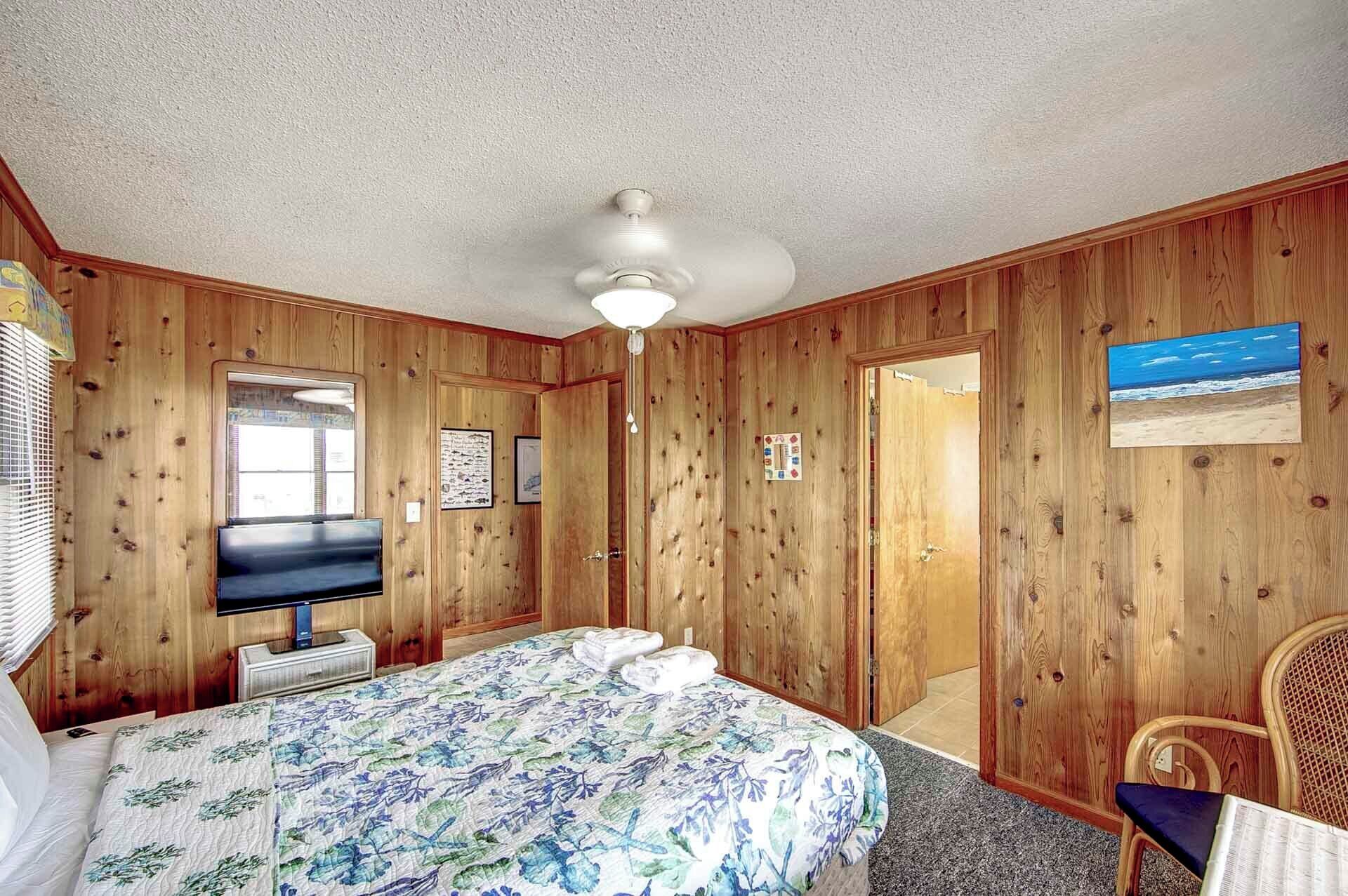 Ein Zimmer mit Holzvertäfelung, einem Bett mit blumigem Bettzeug, einem Deckenventilator, einem Fernseher und einem gerahmten Bild an der Wand.