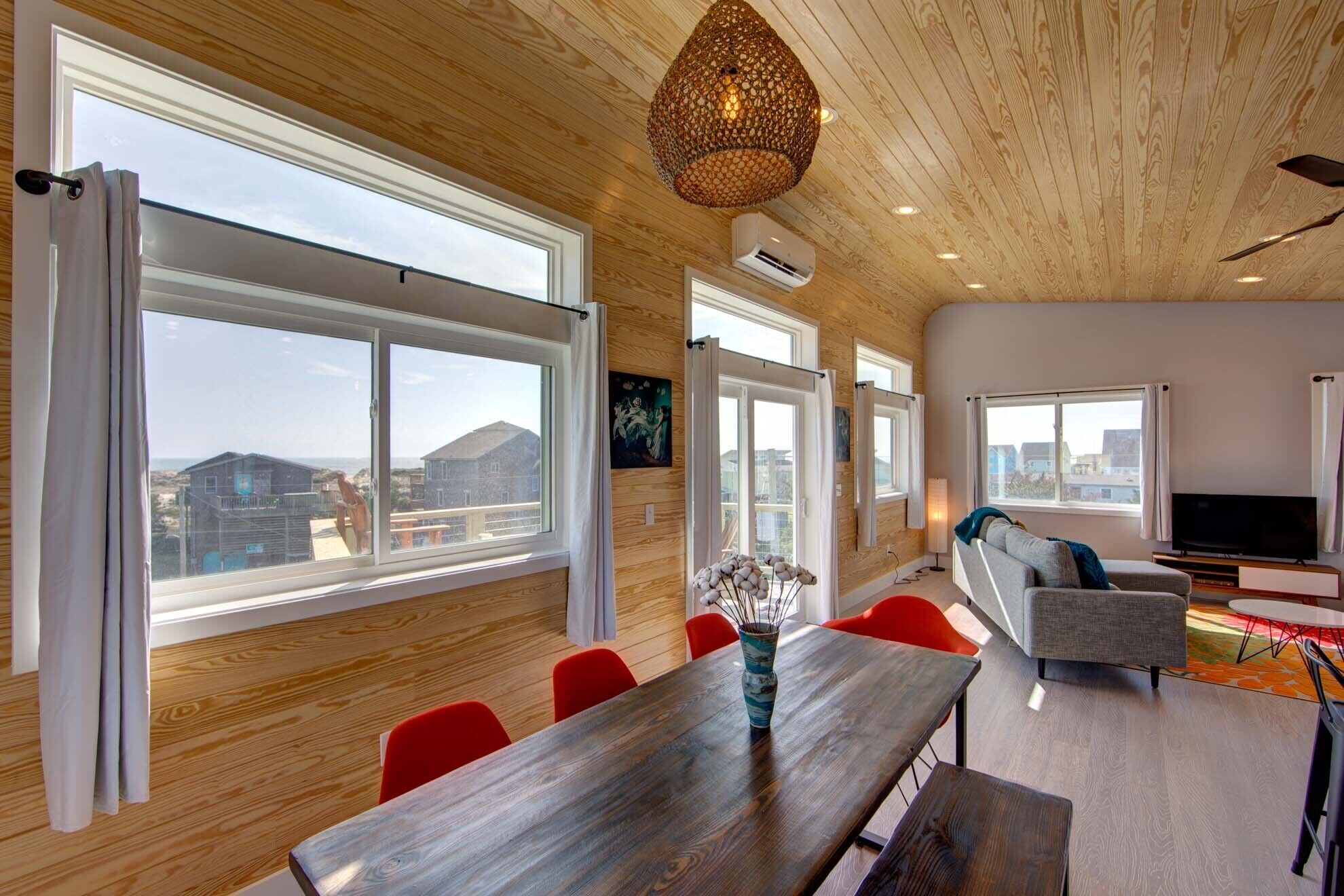 A modern living room with large windows, a wooden ceiling, and a dining area with red chairs.