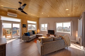 A spacious living room with a wooden ceiling, large windows, a ceiling fan, and a television.