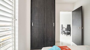 A bedroom with a dark wooden wardrobe, a bed with a red and white patterned bedspread, and a window with white blinds.