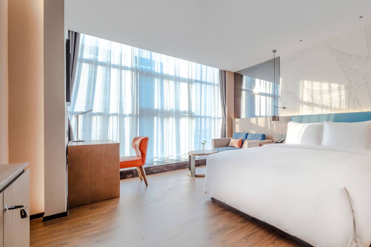 A modern hotel room with a large bed, a desk, and a chair. There is a large window with curtains, a wall-mounted TV, and a decorative wall piece.