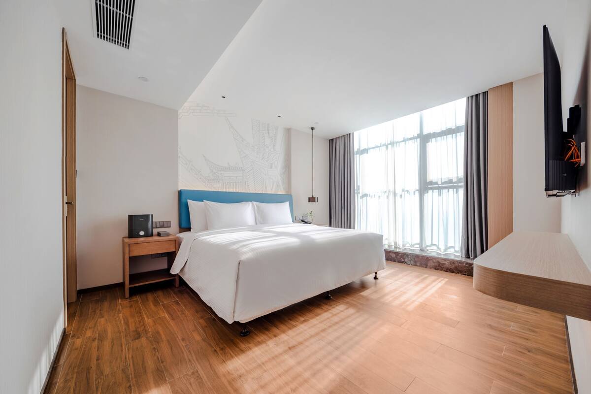 A modern hotel room with a large bed, wooden flooring, a nightstand, a TV, and a window with curtains.