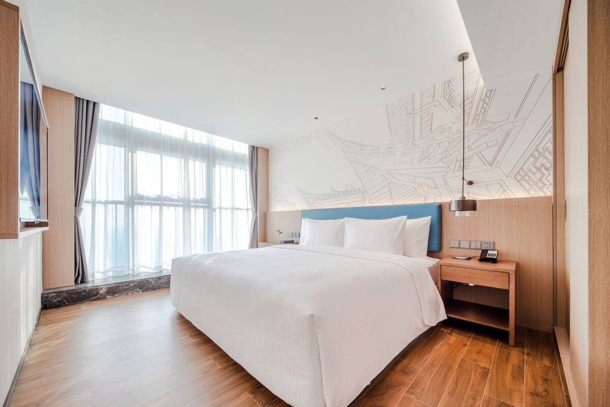 A modern hotel room with a large bed, wooden flooring, a nightstand with a lamp, and a wall with architectural drawings.