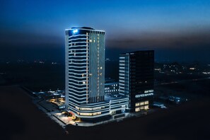 A modern high-rise hotel with a curved facade and illuminated windows, surrounded by a parking area and other buildings. - Shanxi Xiaohe Xincheng Talent Apartment (Taiyuan)