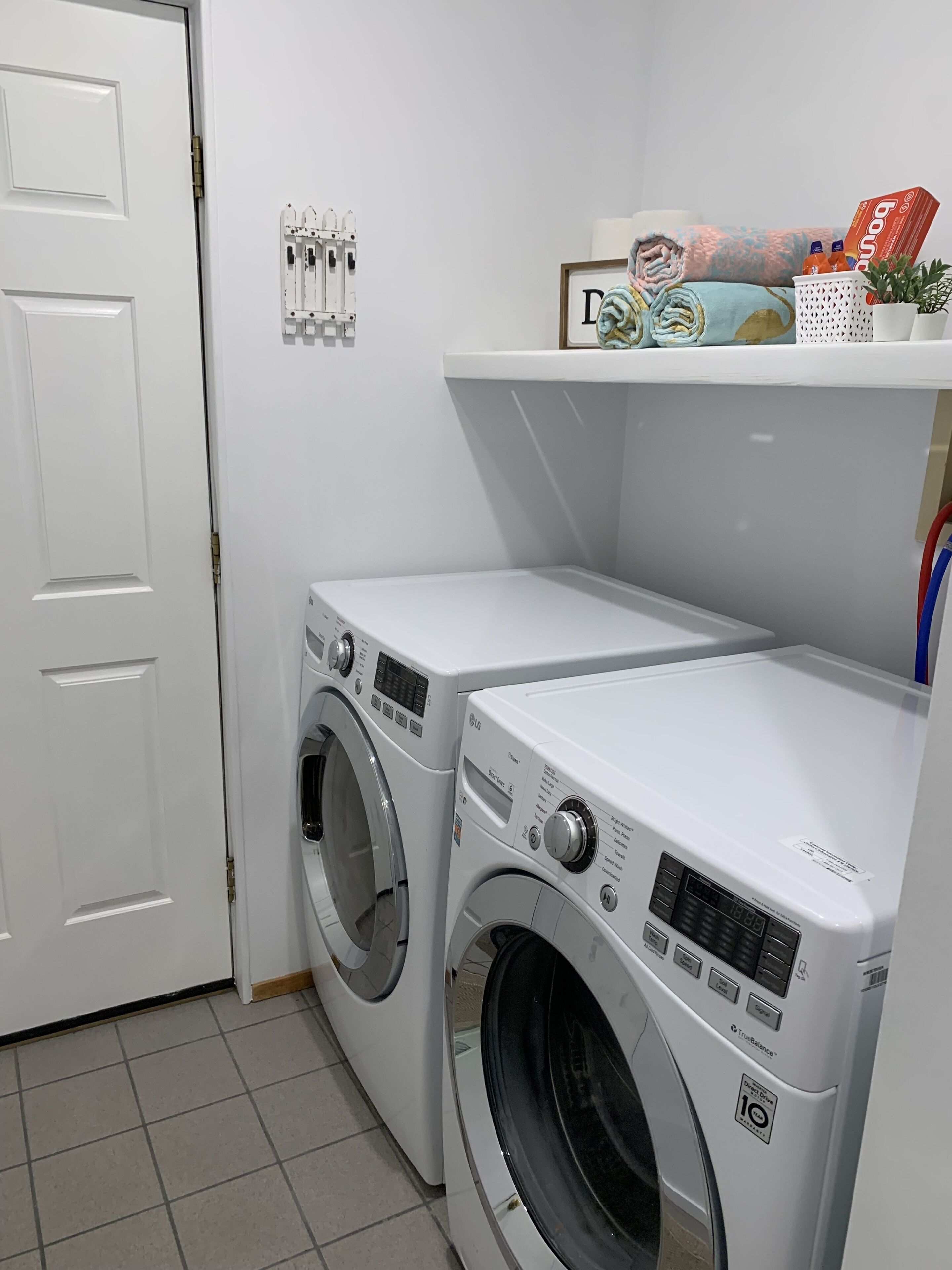 A laundry room with two white front-loading washing machines, a door, and a shelf with a box and bottles.